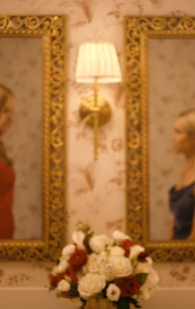 "Two women in elegant dresses stand facing each other in a warmly lit, ornate bathroom with floral wallpaper, gold-framed mirrors, wall lamps, and a vase of flowers on the counter, their reflections visible in the mirrors."