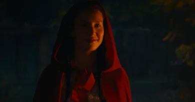 "A young woman wearing a red hooded cloak stands outdoors at night, illuminated softly, with trees and dim lighting in the background, evoking a fairytale atmosphere."
