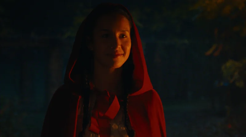 "A young woman wearing a red hooded cloak stands outdoors at night, illuminated softly, with trees and dim lighting in the background, evoking a fairytale atmosphere."