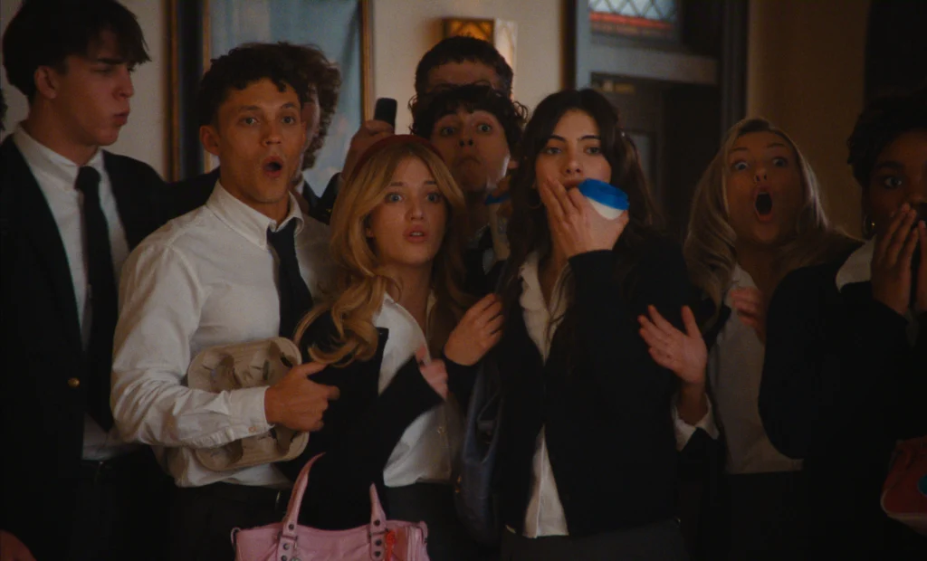 "A group of shocked students in school uniforms stands indoors in a hallway, reacting with surprised expressions as they look at something off-camera."