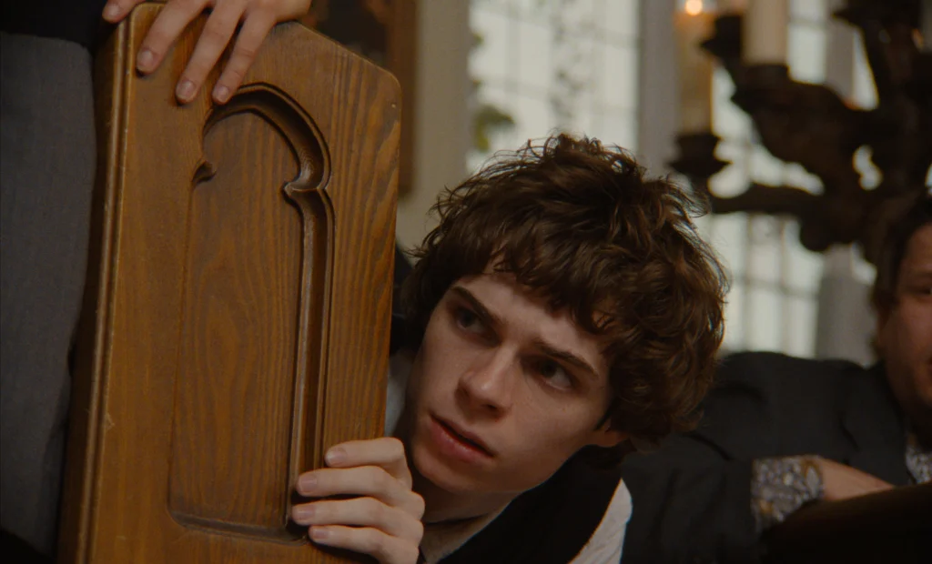 "Young man with curly hair crouches behind a wooden chair, peering cautiously in a dimly lit room with candles and other people in the background, suggesting a tense or suspenseful atmosphere."