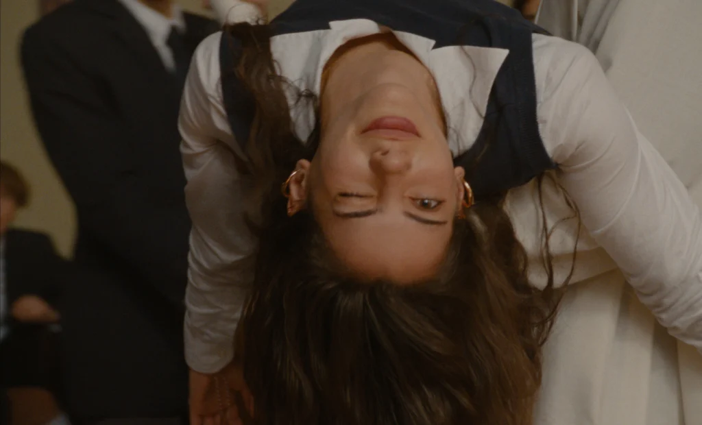 "Young woman in a school uniform is upside down, winking playfully. The indoor setting suggests a formal or academic environment, with other people blurred in the background."