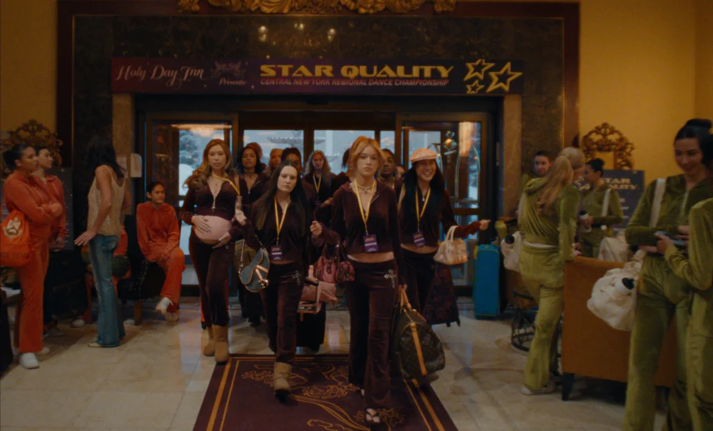 "A group of teenage girls in matching tracksuits walk confidently through a hotel lobby decorated for a regional dance championship, surrounded by other contestants and event signage."